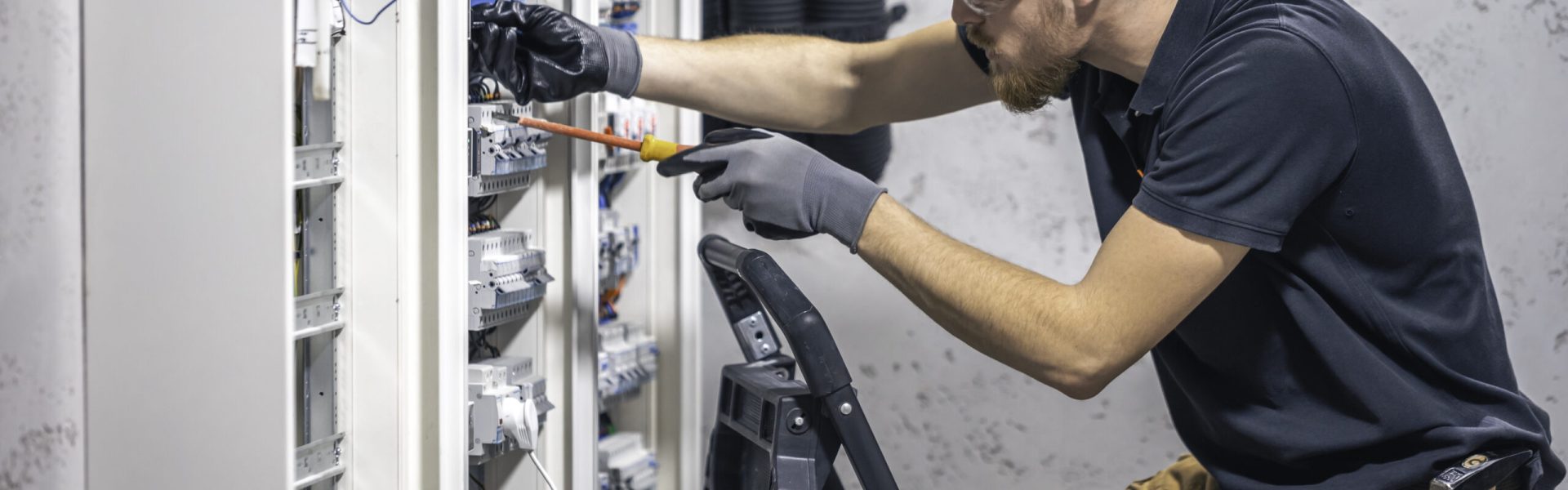 A male electrician works in a switchboard with an electrical connecting cable. Electrician with screwdriver tightens electrically operated switching equipment in fuse box.